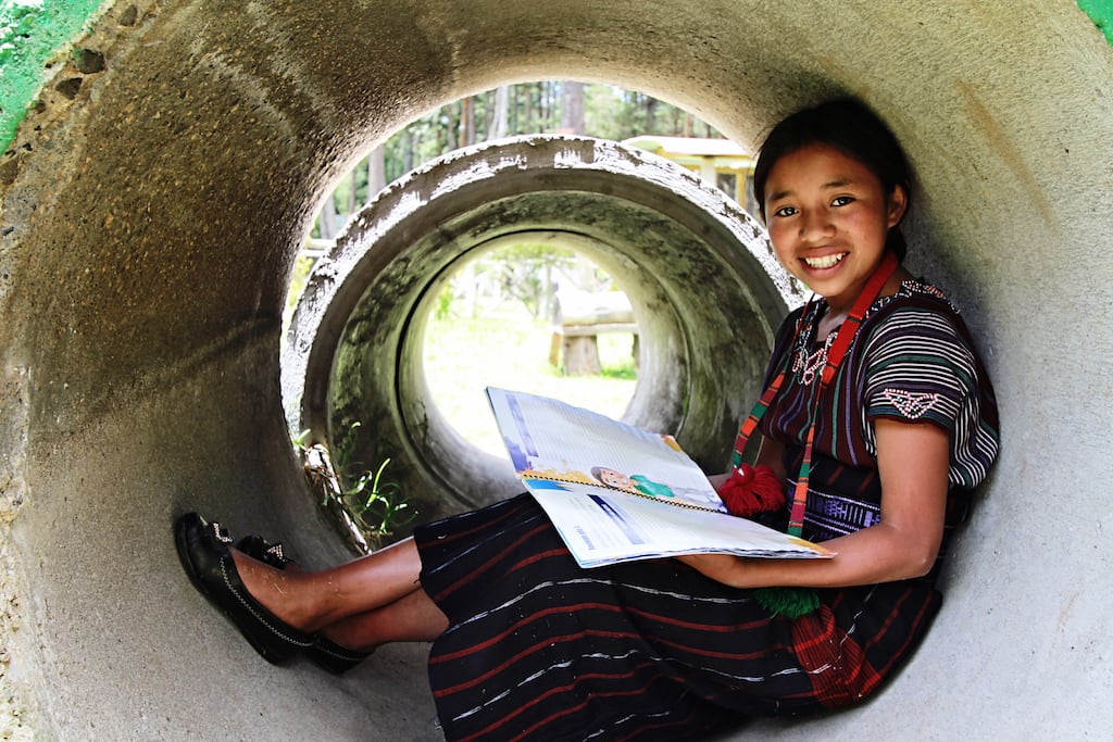 Little girl wearing a red scarf sits in a large concrete tube reading her book, smiling.